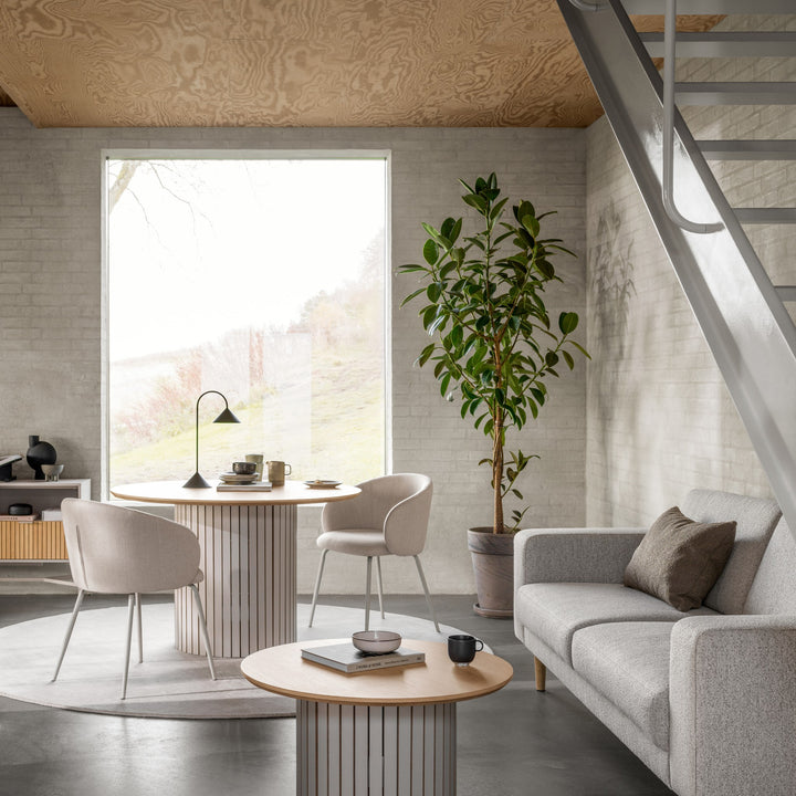 Eleanor spisestol i beige stoff ved rundt søylebord i eik, i stue med grønn plante og grå sofa.