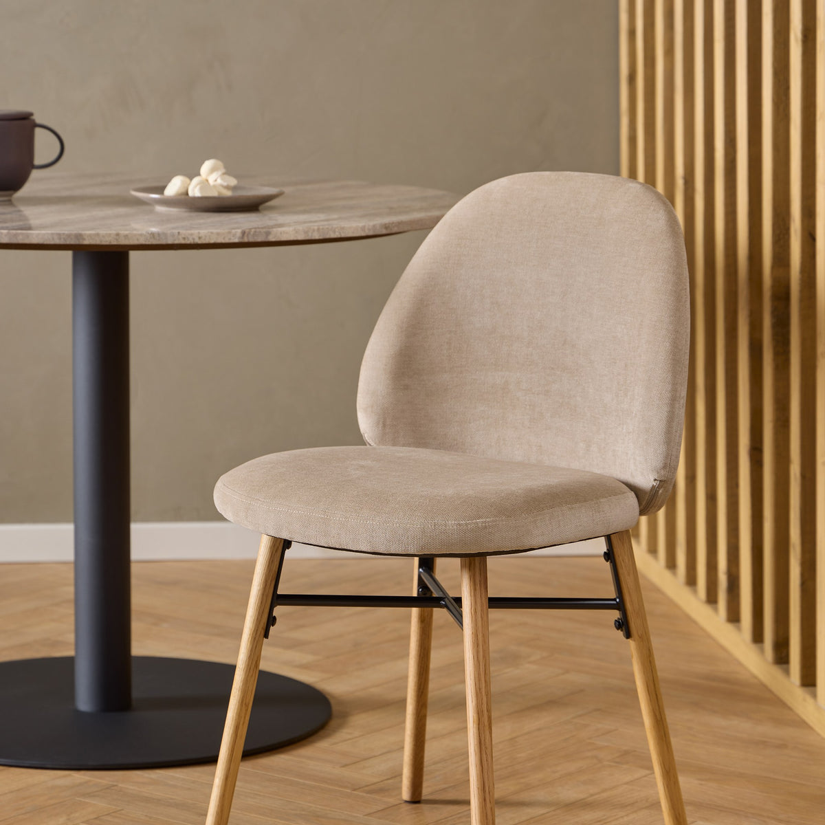 Sophia spisestol i beige velur med eikeben, ved rundt steinbord foran trespilevegg