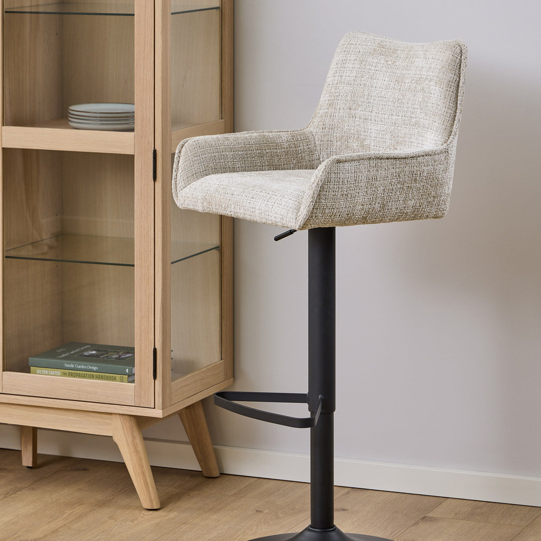 Eddie barstol i beige chenille med armlener og sort fot, ved eikevitrine med bøker