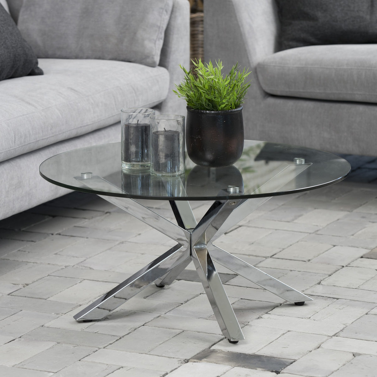 Heaven sofabord med klar glassplate og kromben foran grå sofa, med plante