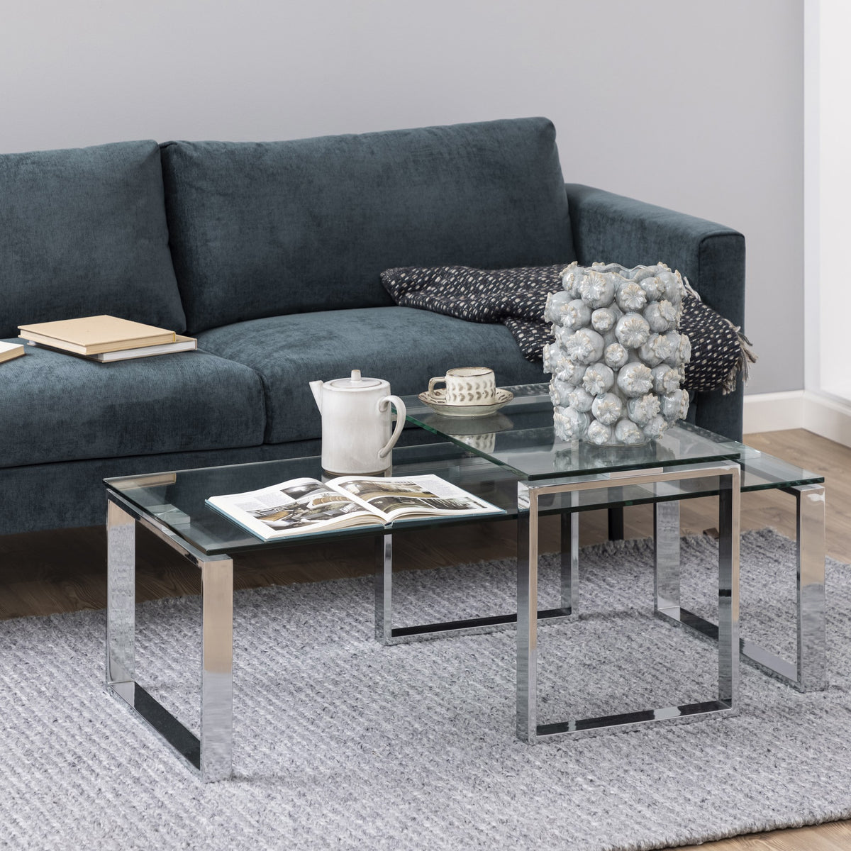 Katrine sofabord, sett med klare glassplater og kromben foran mørk grå sofa