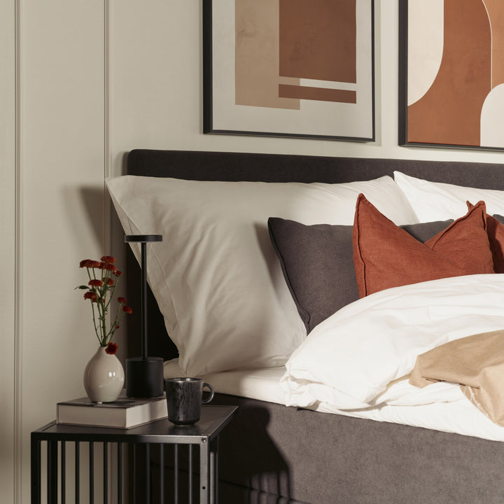 Strington sidebord i sort metall med vase, lampe og kopp ved seng med terrakottapute