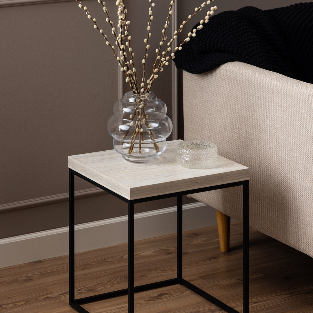 Barossa sidebord i lys travertinlook med sort metallramme, ved seng med vase og pledd