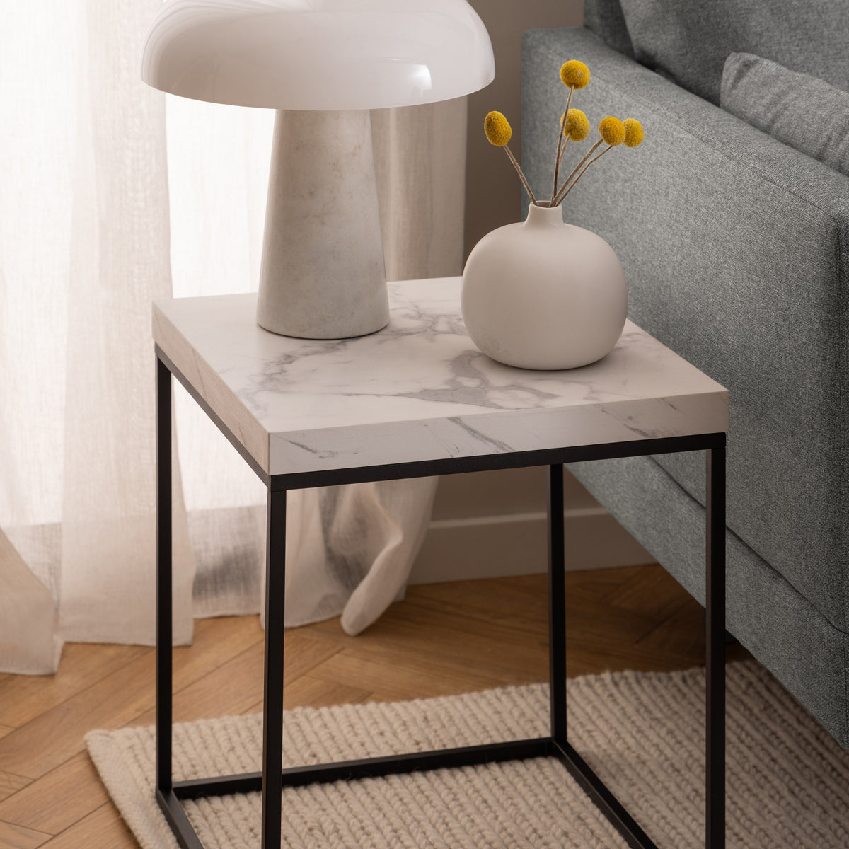 Barossa sidebord i hvit marmorlook med sort metallramme, ved grå sofa med lampe og vase