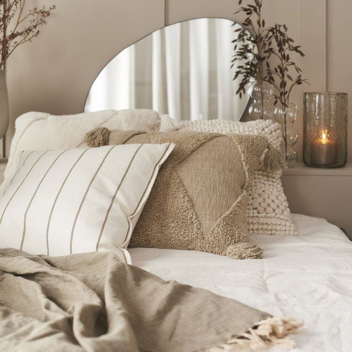 Hill pyntepute i offwhite med smale striper, på en seng med beige tuftet pute og lys