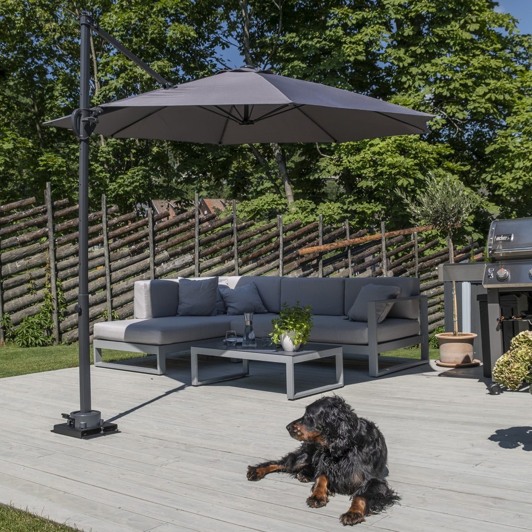 Smart parasollfot i sort med grå hengeparasoll over grå loungesofa på terrasse, hund i hagen.