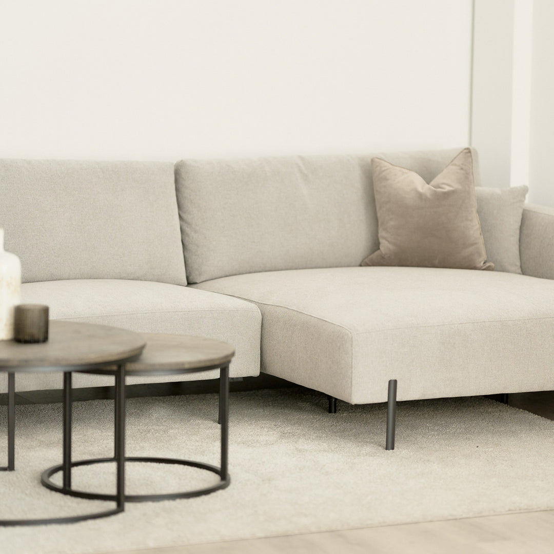 Milazzo hjørnesofa i lys beige stoff med pyntepute i lys stue, runde settebord i mørkt tre foran