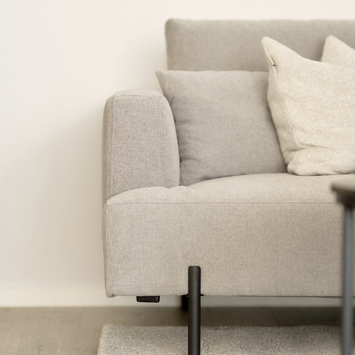 Milazzo hjørnesofa i nærbilde med lys beige stoff, avrundet armlene og pyntepute mot lys vegg