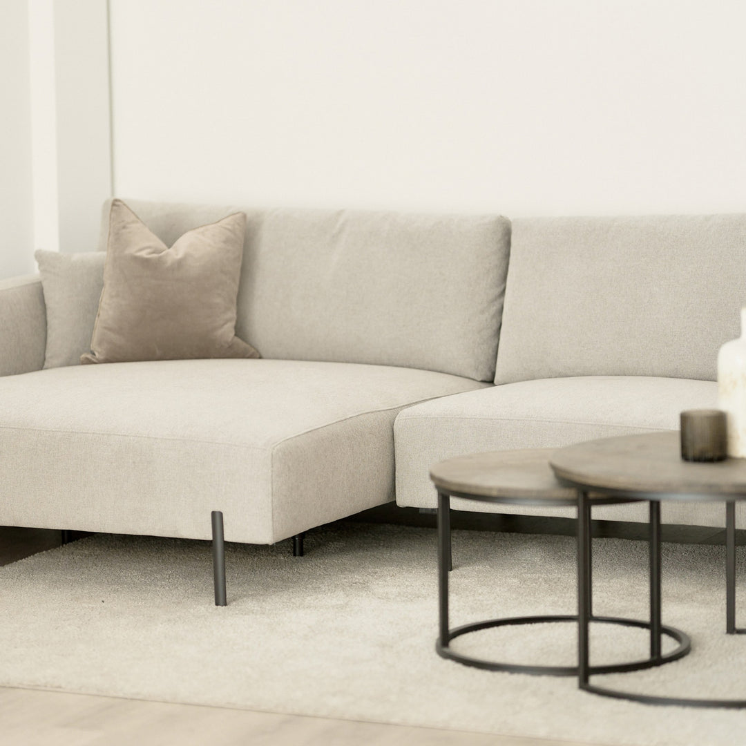 Milazzo hjørnesofa i lys beige stoff med beige pyntepute i lys stue, runde settebord i mørkt tre foran