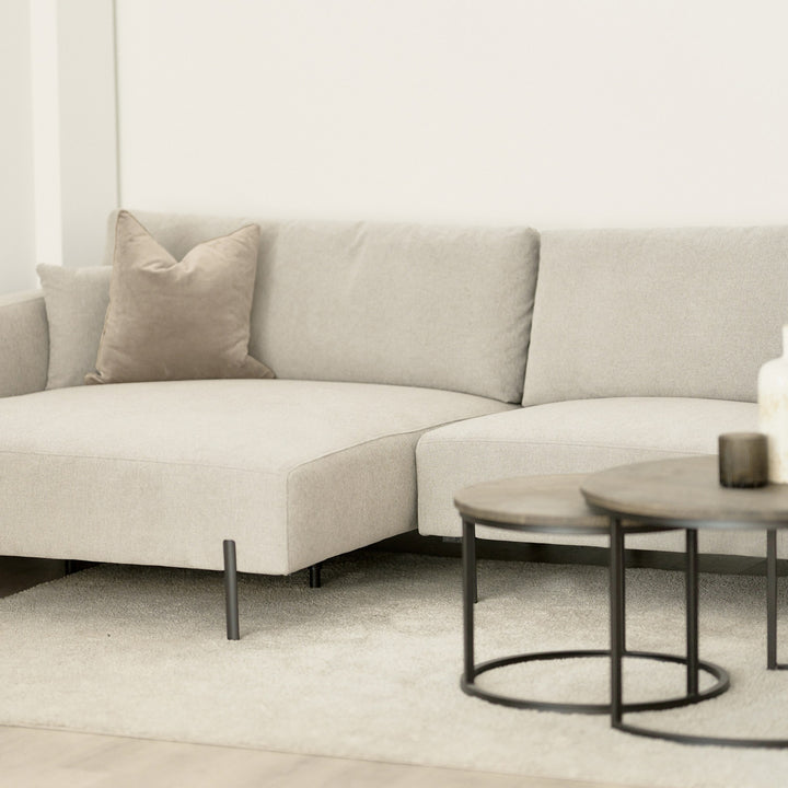 Milazzo hjørnesofa i lys beige stoff med beige pyntepute i lys stue, runde settebord i mørkt tre foran