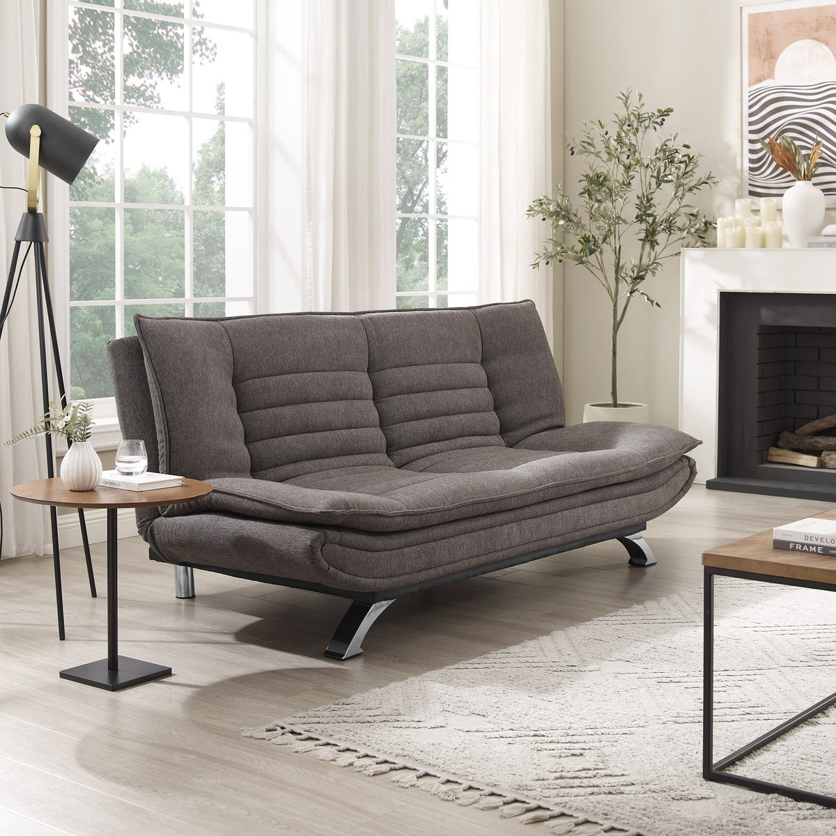 Friday sovesofa i mørk grå stoff, i lys stue med peis, kunstig tre og stativlampe