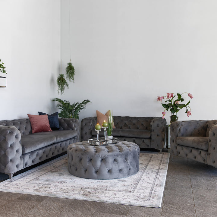 Bella sofa i grå tuftet velur med rund puff og lenestol, lyst rom med planter og blomster.