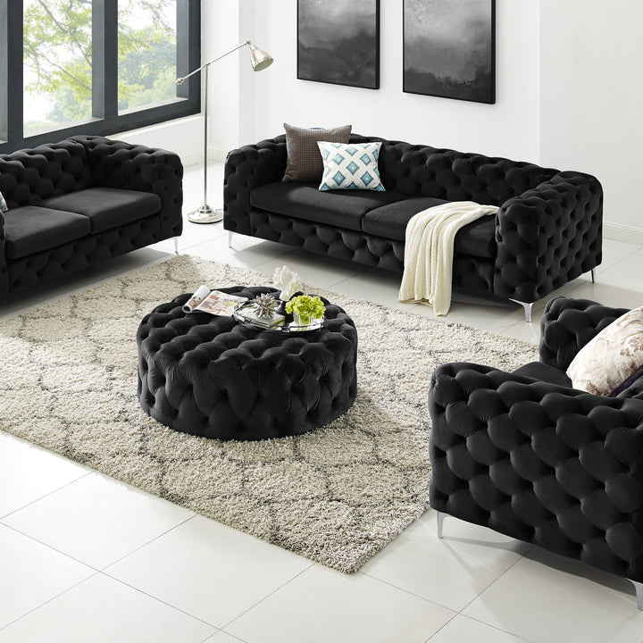 Bella sofa i sort velur, tuftet, med rund puff og lenestol i lys stue med mønstret teppe og bilder