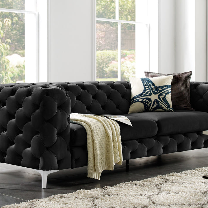 Bella sofa i sort velur med knappetrekk, med pledd og puter, i lys stue med store vinduer og shaggteppe