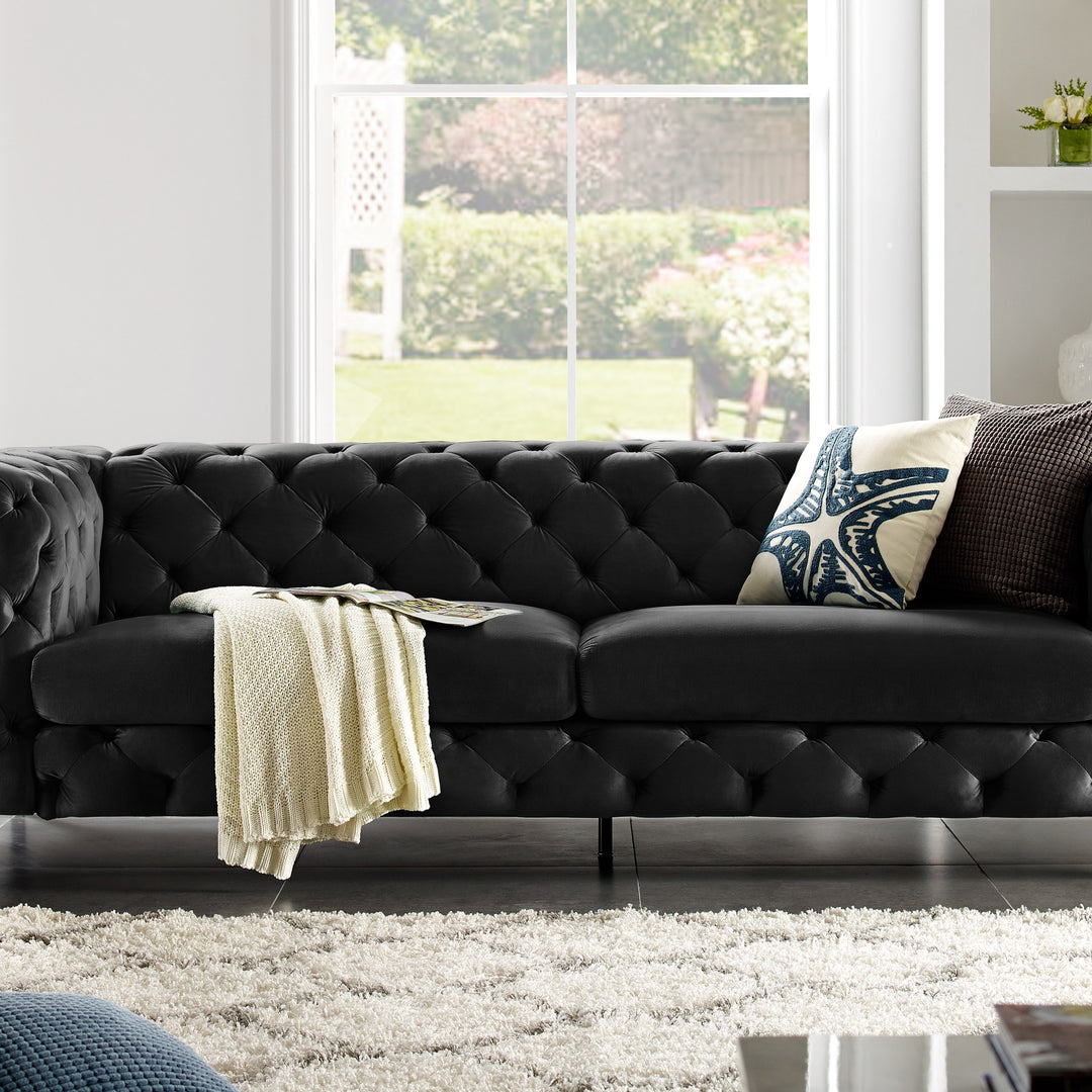 Bella sofa i sort tuftet velur, lyst stuerom med strikkepledd, puter og hvitt ryeteppe.