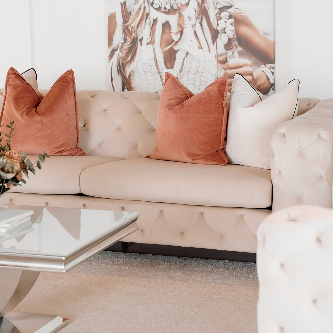Bella sofa i krem velur, treseter med terrakotta puter, glassbord og boho veggbilde i lys stue