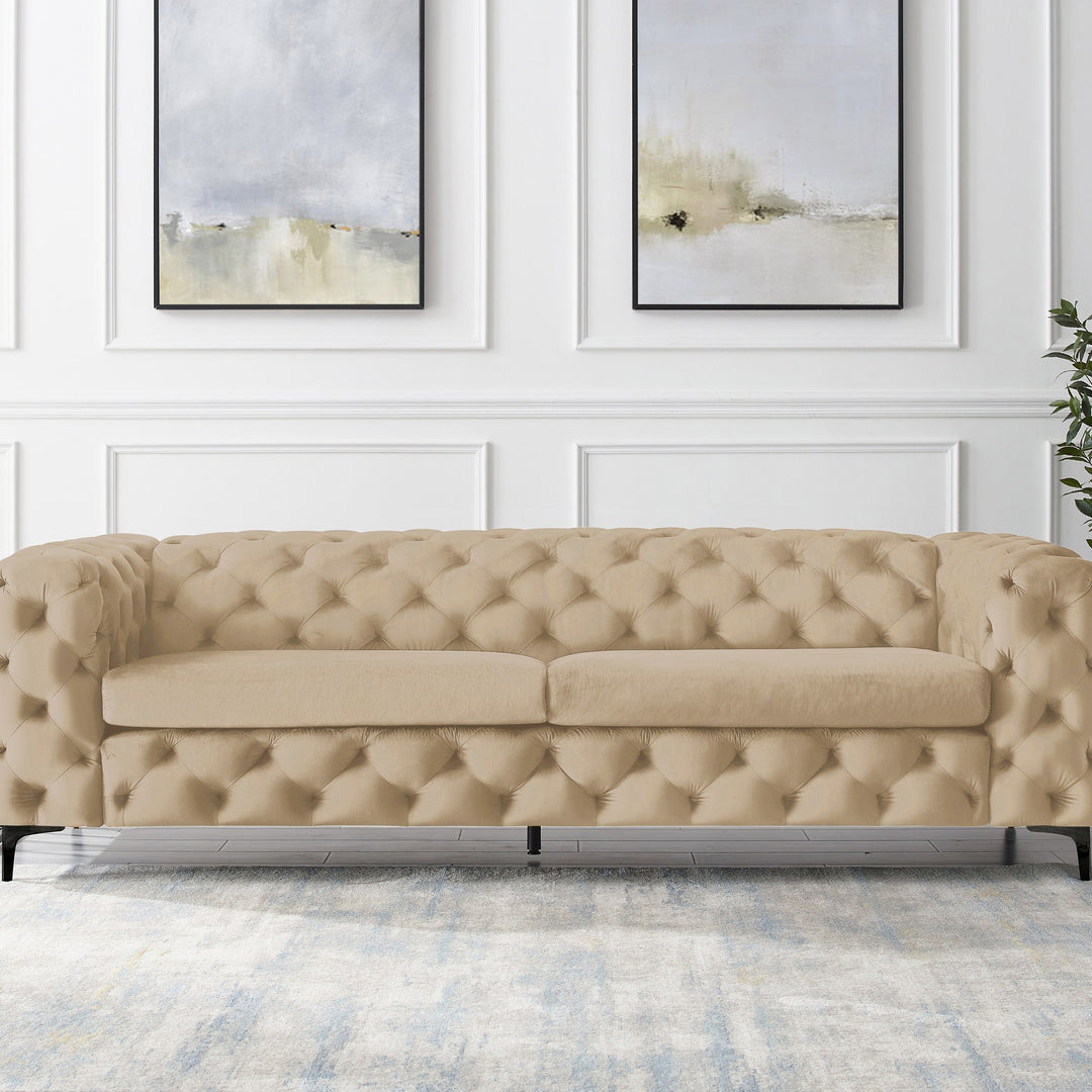 Bella sofa i beige tuftet velur, panelvegg med abstrakt kunst og grønn plante.