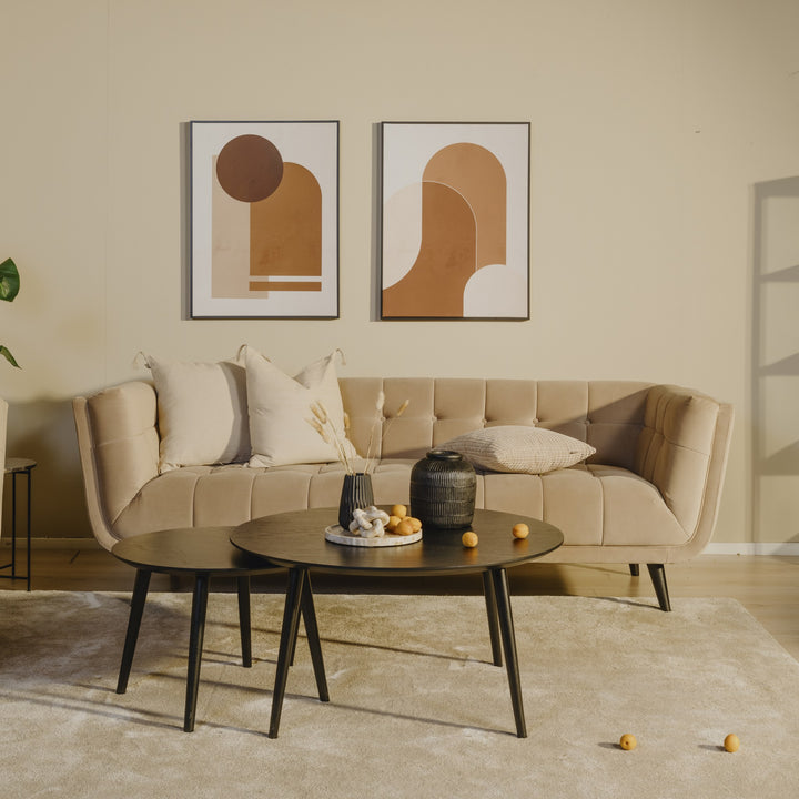 Simon sofa i beige velur i varm stue med abstrakte bilder, sorte sofabord og grønn plante.
