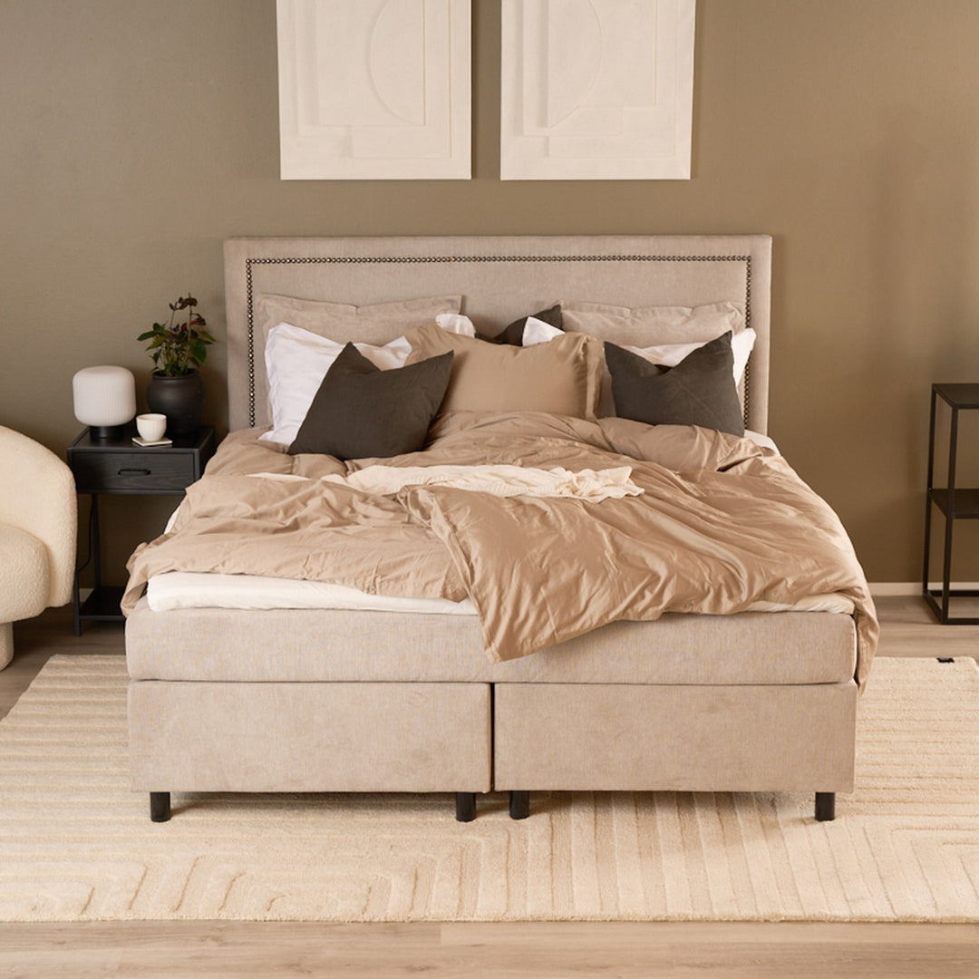 Saturn Lux kontinentalseng i beige med pynteputer, nattbord med plante og lyst teppe