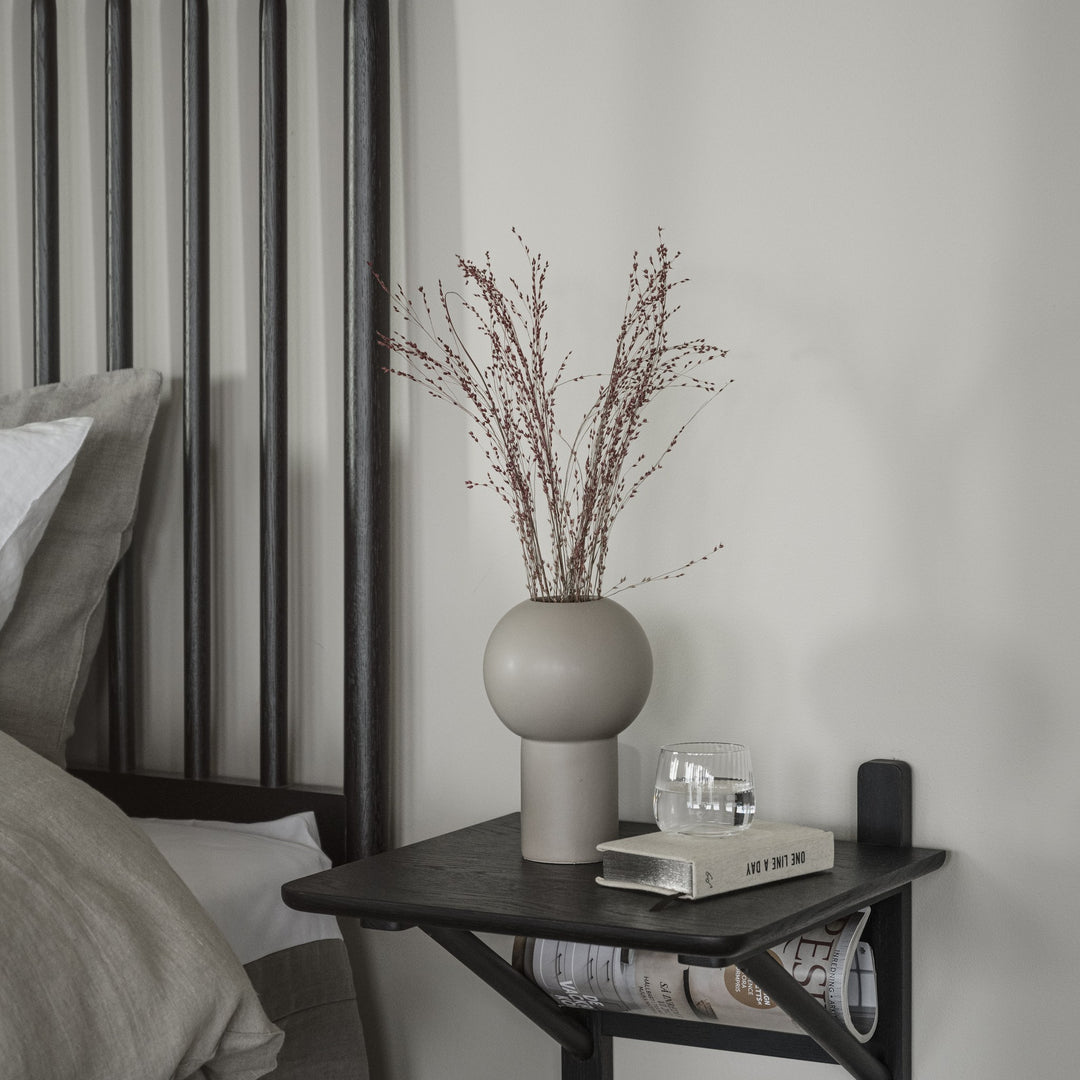 Rowico Riggins sengegavl i sort eik med sort natthylle, beige vase med tørkede blomster og bok