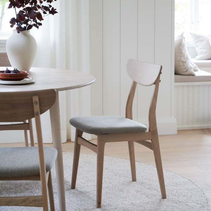 Rowico Rodham spisestol i hvitpigmentert eik med gråbeige sete, ved rundt spisebord i lyst rom.