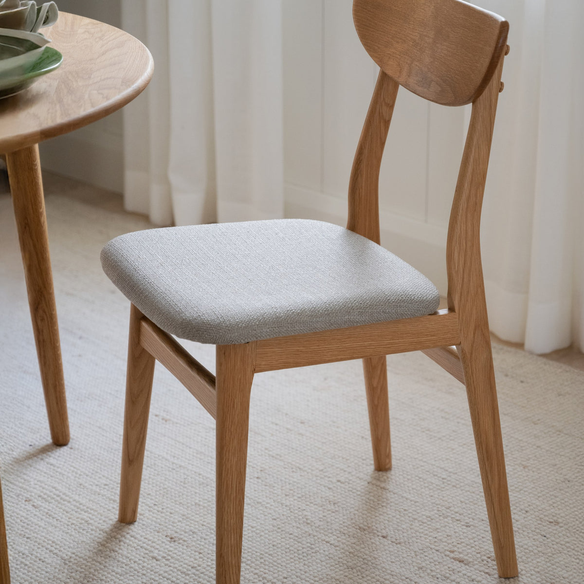 Rowico Rodham spisestol i eik med gråbeige stoffsete, ved rundt eikebord på beige teppe i lyst rom