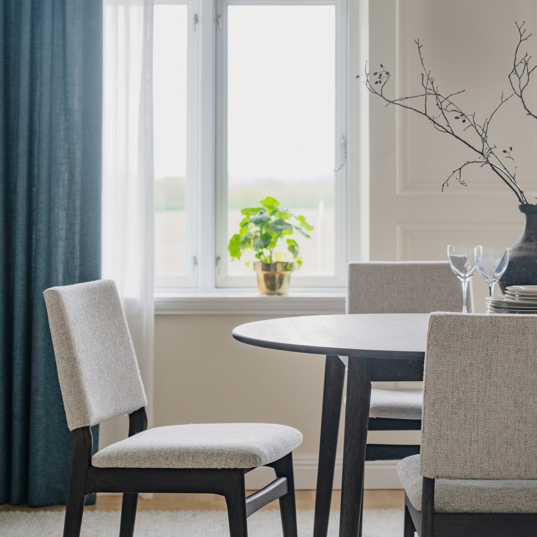Rowico Medford spisestol i sortbeiset eik med lyst sete ved svart rundbord og blå gardin
