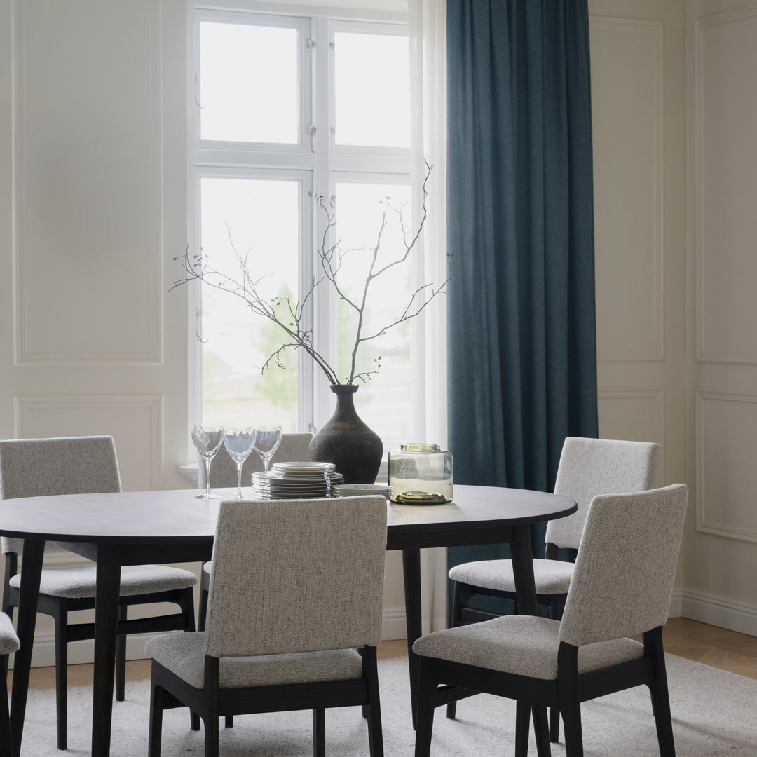 Rowico Medford spisestol i sortbeiset eik med lyst sete ved svart ovalt bord og blå gardin