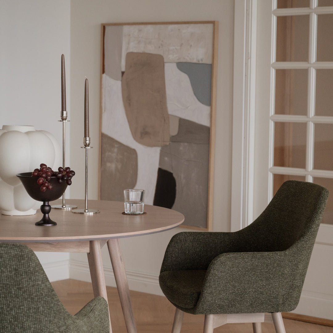 Rowico Denley spisestol i mørkegrønn stoff med armlener ved lyst rundt eikebord med kunst