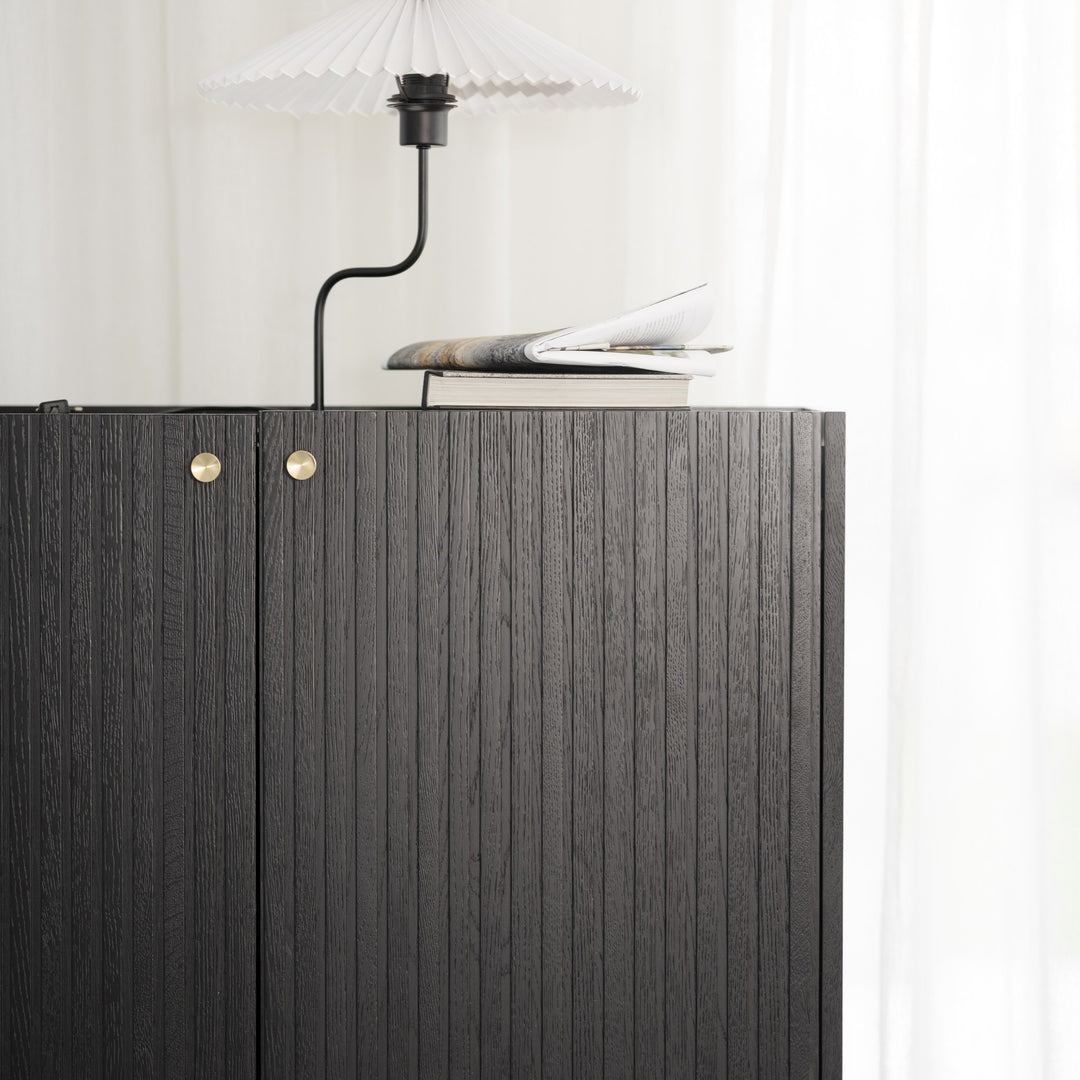 Rowico Halifax skap i sort rillet eik med plissert lampe og bok på toppen, ved lyst gardin