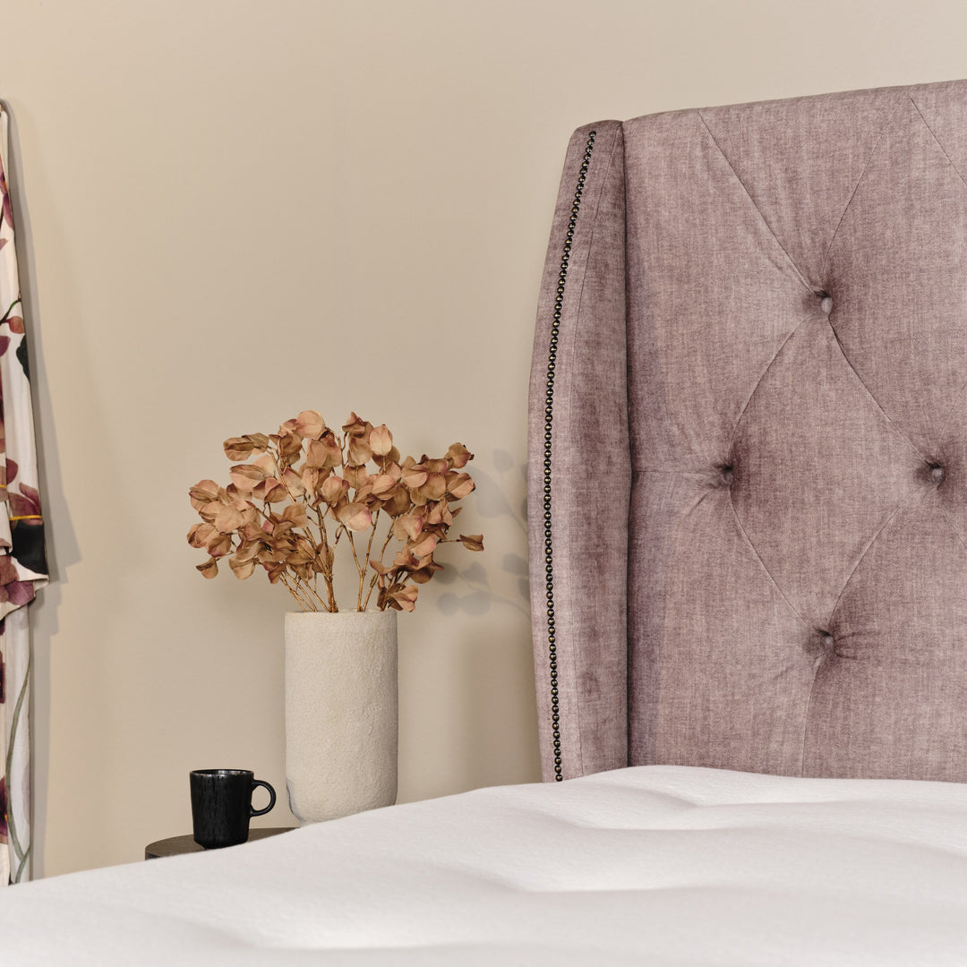 Asleep Bellamy sengegavl i dempet rosa ved beige vegg, med vase med tørkede blomster og svart kopp
