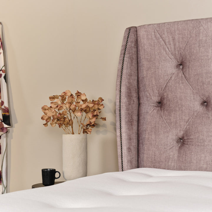 Asleep Bellamy sengegavl i dempet rosa ved beige vegg, med vase med tørkede blomster og svart kopp