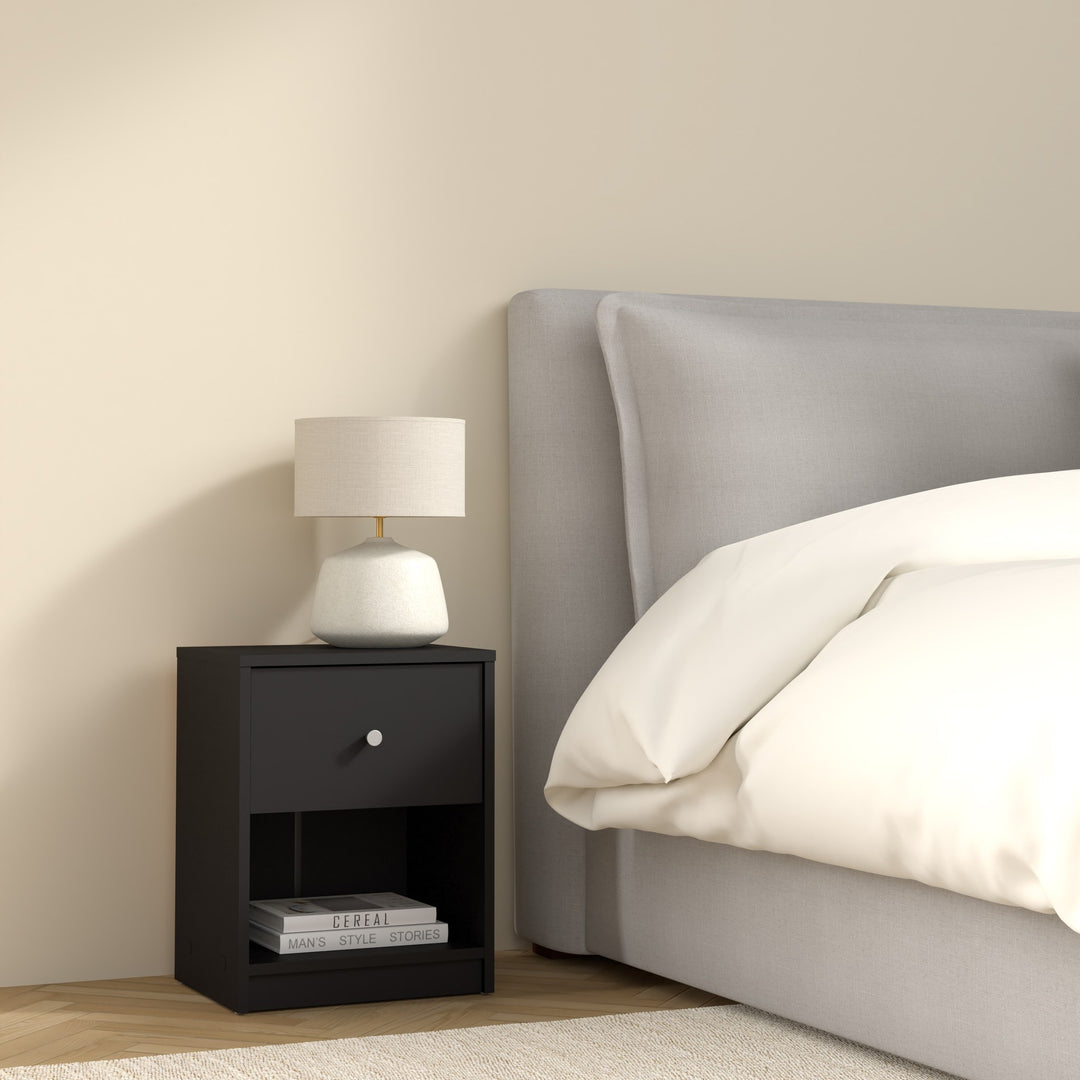 May nattbord i sort med skuff og åpen hylle, med lampe ved grå seng i soverom