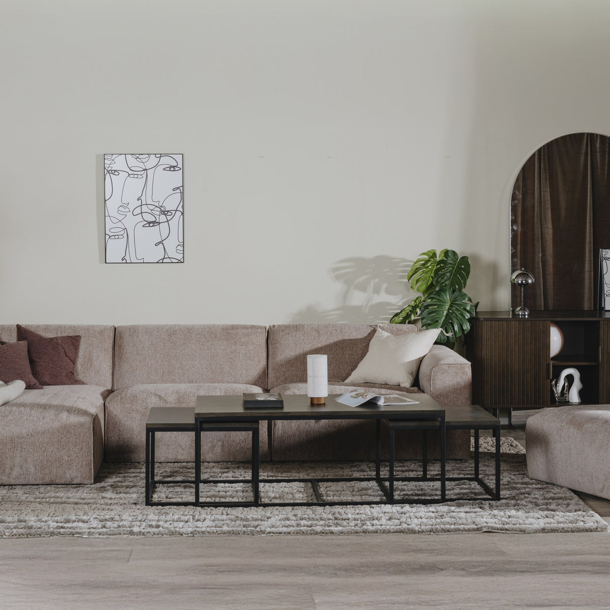 Como hjørnesofa i gråbrun stoff med sjeselong, sorte sofabord og buet veggspeil i lyst rom