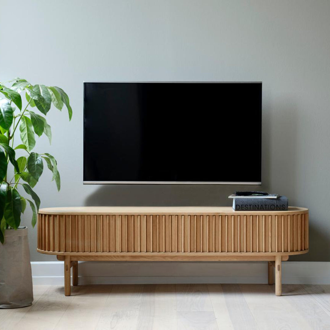 Carno TV-benk i eik med rillet front og runde ben under vegghengt TV med plante