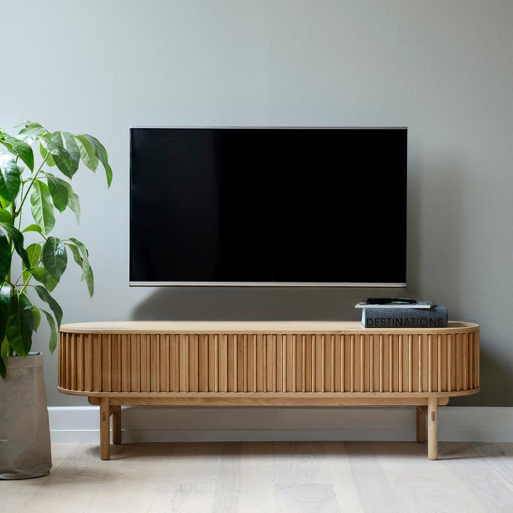 Carno TV-benk i eik med rillet front og runde ben under vegghengt TV med plante