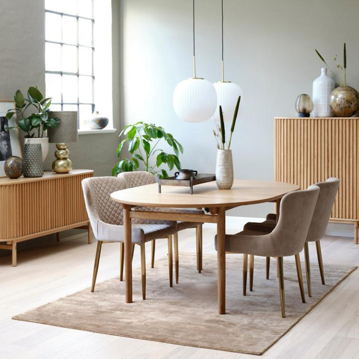 Carno spisebord i eik med beige velurstoler i lyst spiserom med riflede skjenker og grønne planter
