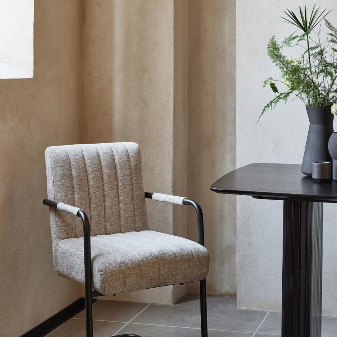 Trout spisestol i sandfarget stoff med svart metallramme og armlener, ved sort bord mot beige vegg