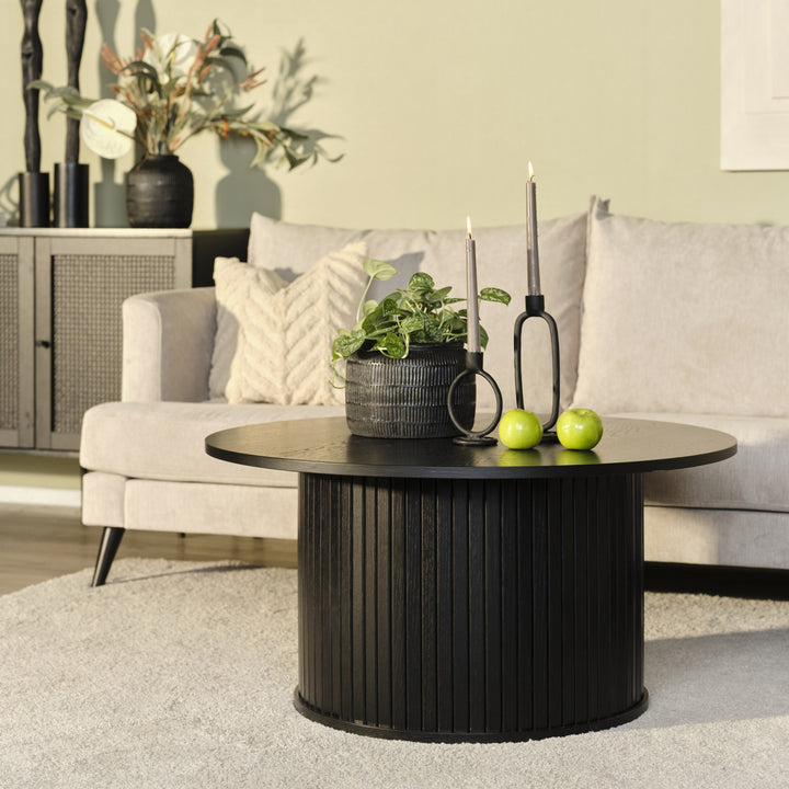 Fiola sofabord i sort eik, rundt med riflet søyle, foran beige sofa med plante og lysestaker