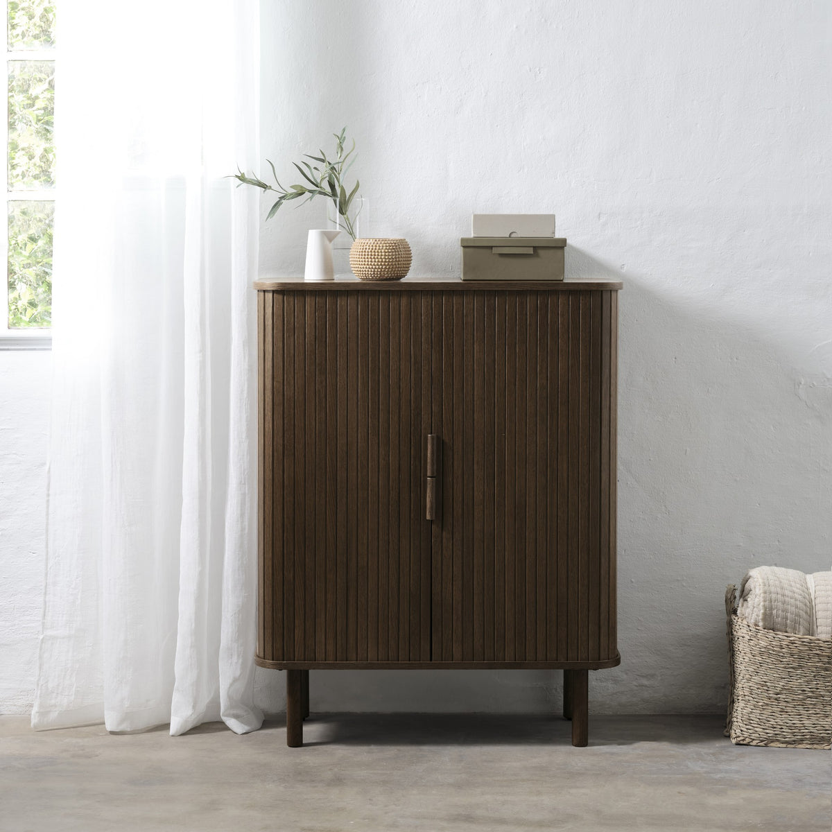 Cavo skap i røyket eik med vertikale spiletredører, mot hvit murvegg med vindu og flettet kurv
