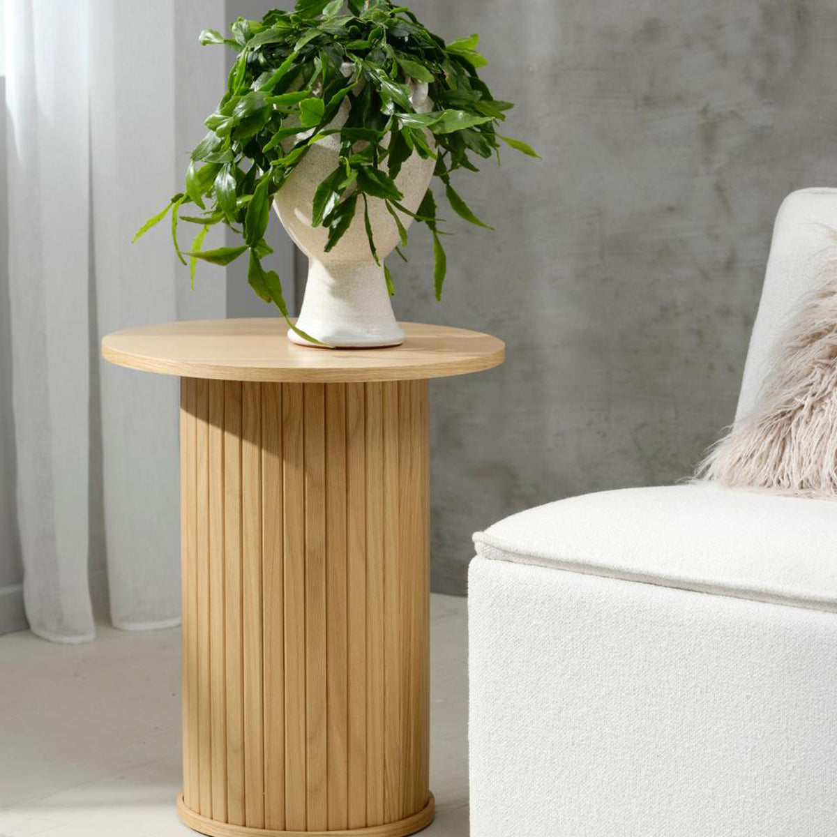 Fiola sidebord i natur eik, rundt med riflet søylefot, ved hvit boucléstol med plante i krukke