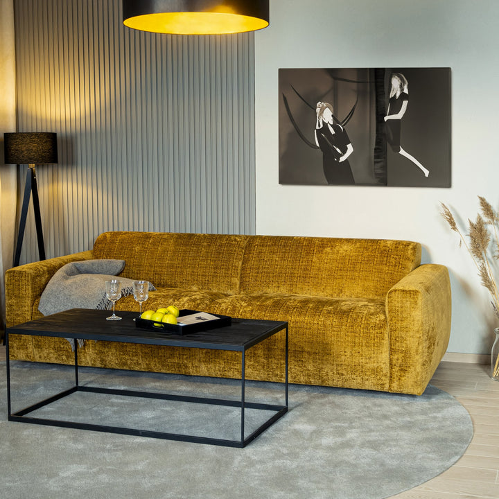 Bellus Olos 3-seter sofa i sennepsgul velur mot grå riflet vegg med sort gulvlampe og rundt sofabord