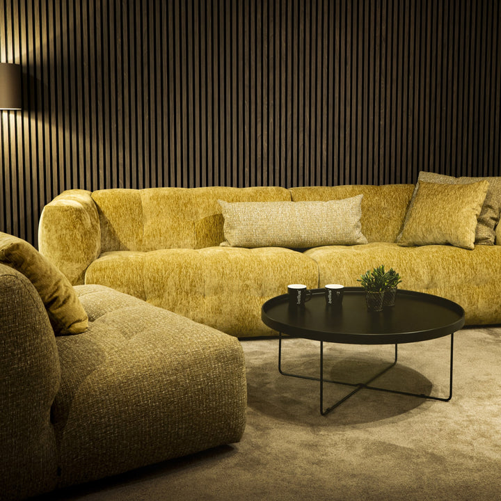 Bellus Soffy sofa med puff i okergul chenille, mørk spilevegg, svart sofabord og grønn plante