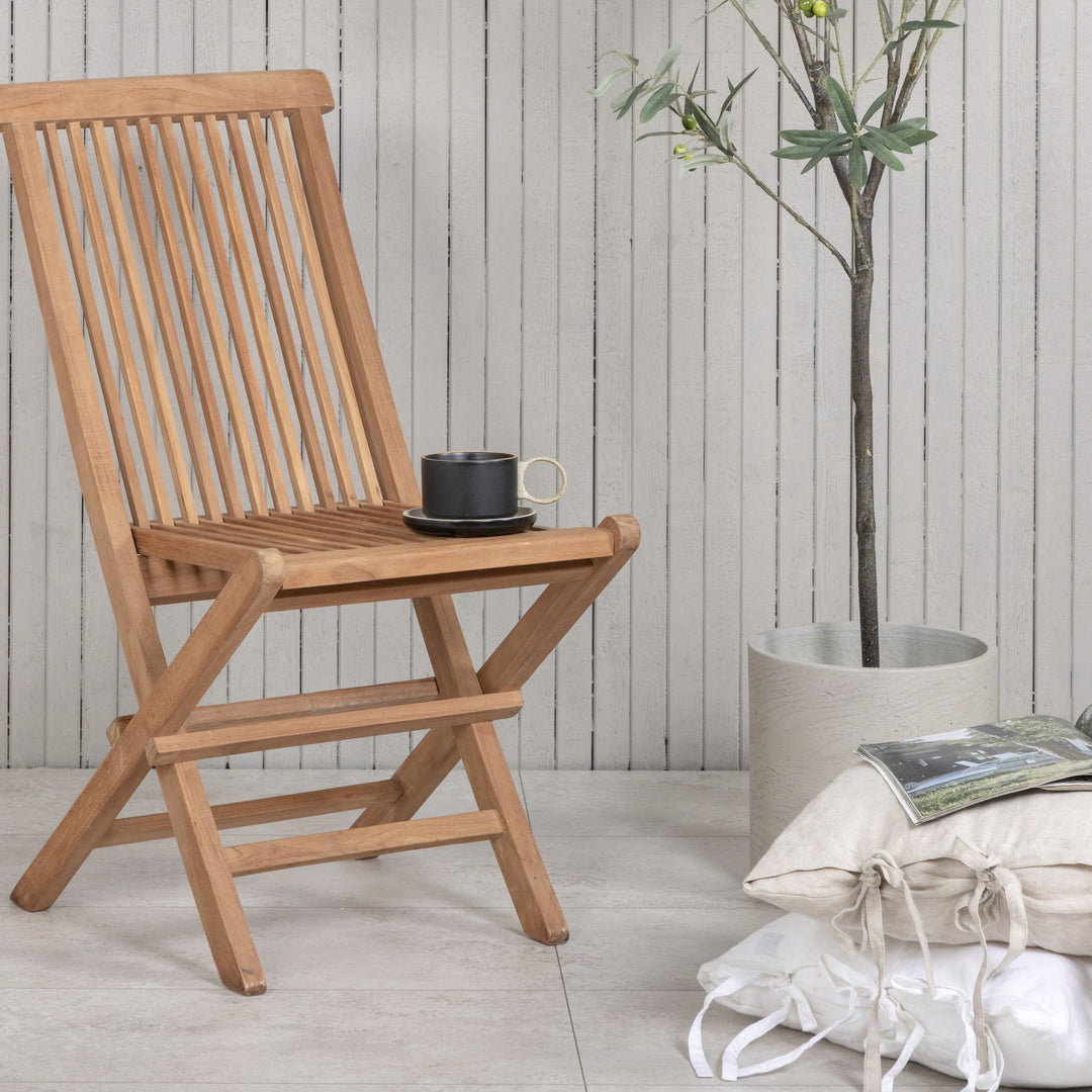 Venture Design Kenya klappstol i natur teak, mot hvit panelvegg med plante og sort kopp på setet