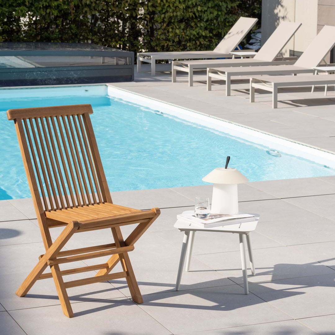 Venture Design Kenya klappstol i teak ved basseng, med solsenger og hvitt sidebord i sollys.
