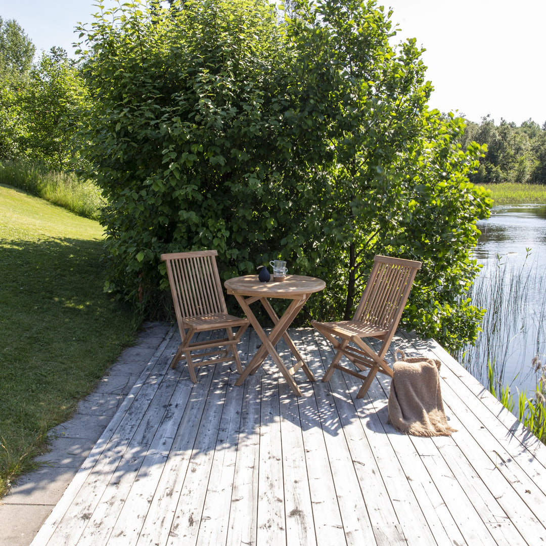 Venture Design Kenya cafébord i natur teak med to klappstoler på trebrygge ved vann og grønt