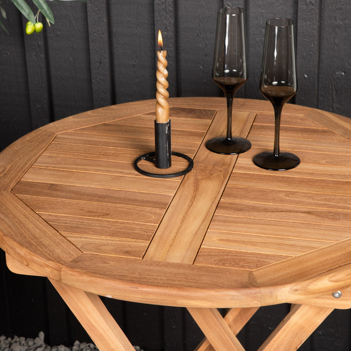 Venture Design Kenya cafébord i teak med levende lys og champagneglass, mot mørk panelvegg ute.