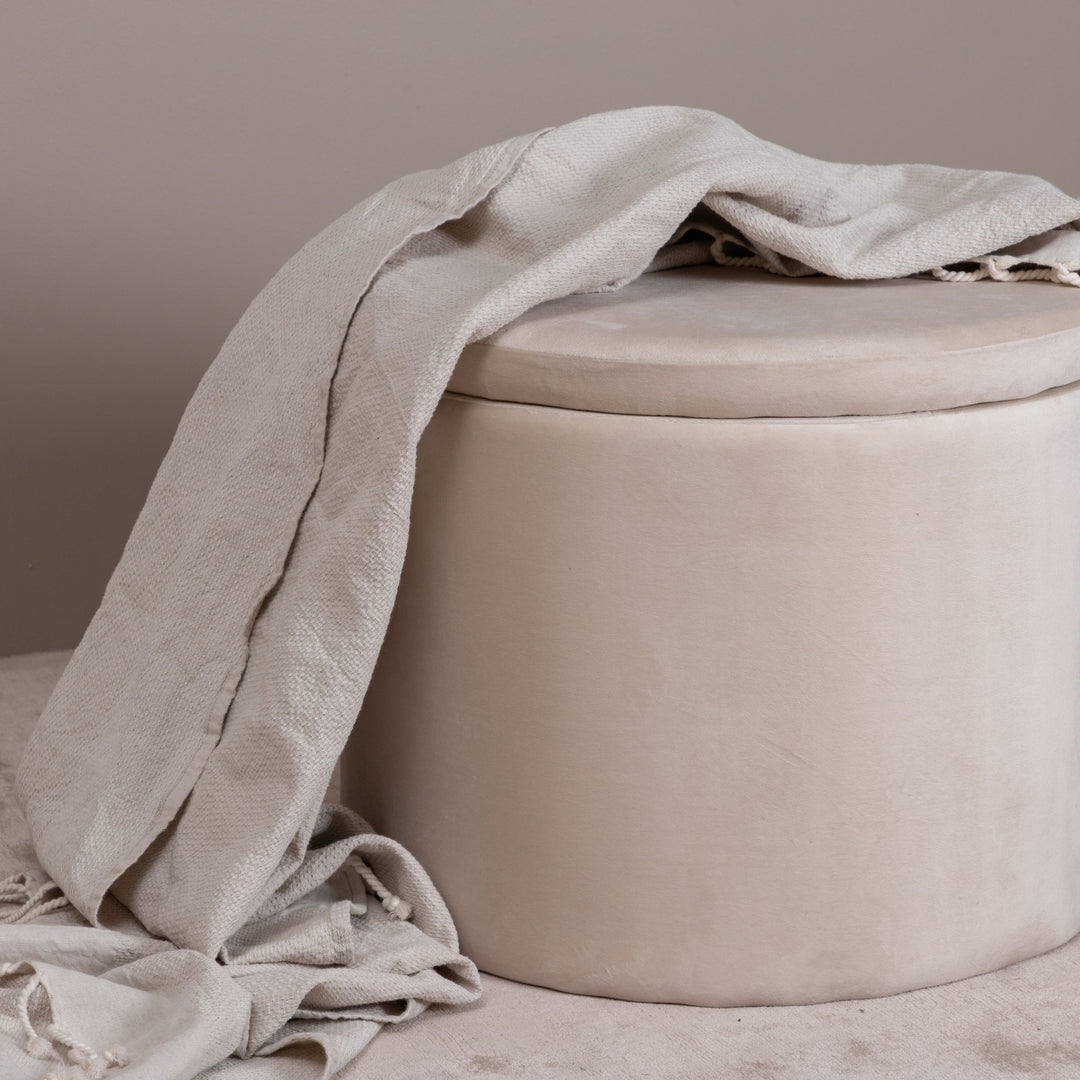 Dunken puff i beige fløyel med lyst pledd drapert over, mot beige vegg