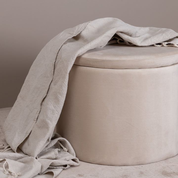 Dunken puff i beige fløyel med lyst pledd drapert over, mot beige vegg