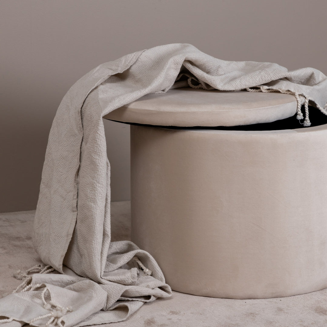 Dunken oppbevaringspuff i beige velur med lyst pledd drapert over og lokket på gløtt.